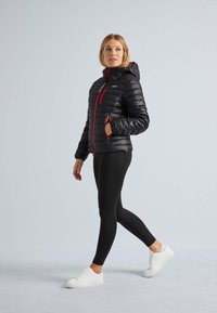 Sort pufferjakke med hætte, der har en rød lynlås, sidelommer og et quiltet mønster. Modellen bærer sorte leggings og hvide sneakers.
