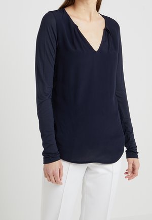 Frau trägt ein eng anliegendes marineblaues Langarm-V-Ausschnitt-Oberteil und weiße Hosen, stehend vor einem einfarbigen hellen Hintergrund.