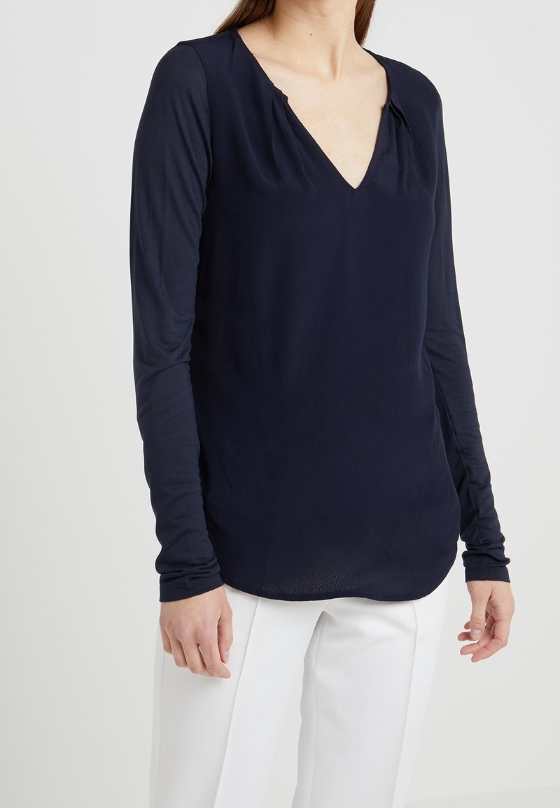 Mujer que lleva una camiseta azul marino ajustada de manga larga con escote en V y pantalones blancos, de pie frente a un fondo liso y claro.
