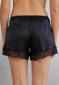 Zwarte satijnen shorts met een scalloped kantzoom. Voorzien van een elastische tailleband. Smooth textuur, met kantdetails aan de randen.