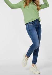 Grön hoodie med dragsnören; tight blå jeans med detaljer på framsidan; kraftiga vita sneakers; avslappnad outfit som passar för vardagligt bruk.