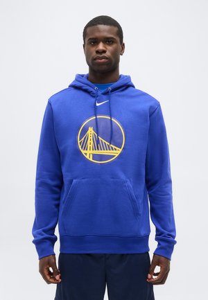 Egy férfi kék kapucnis pulóverben, amelyen sárga híd logó és fehér Nike szimbólum látható, világos, egyszínű háttér előtt áll.