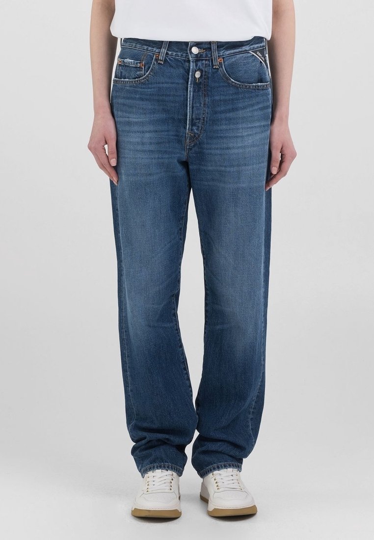 Personne portant un jean bleu droit taille haute et des baskets blanches, debout devant un fond uni clair.
