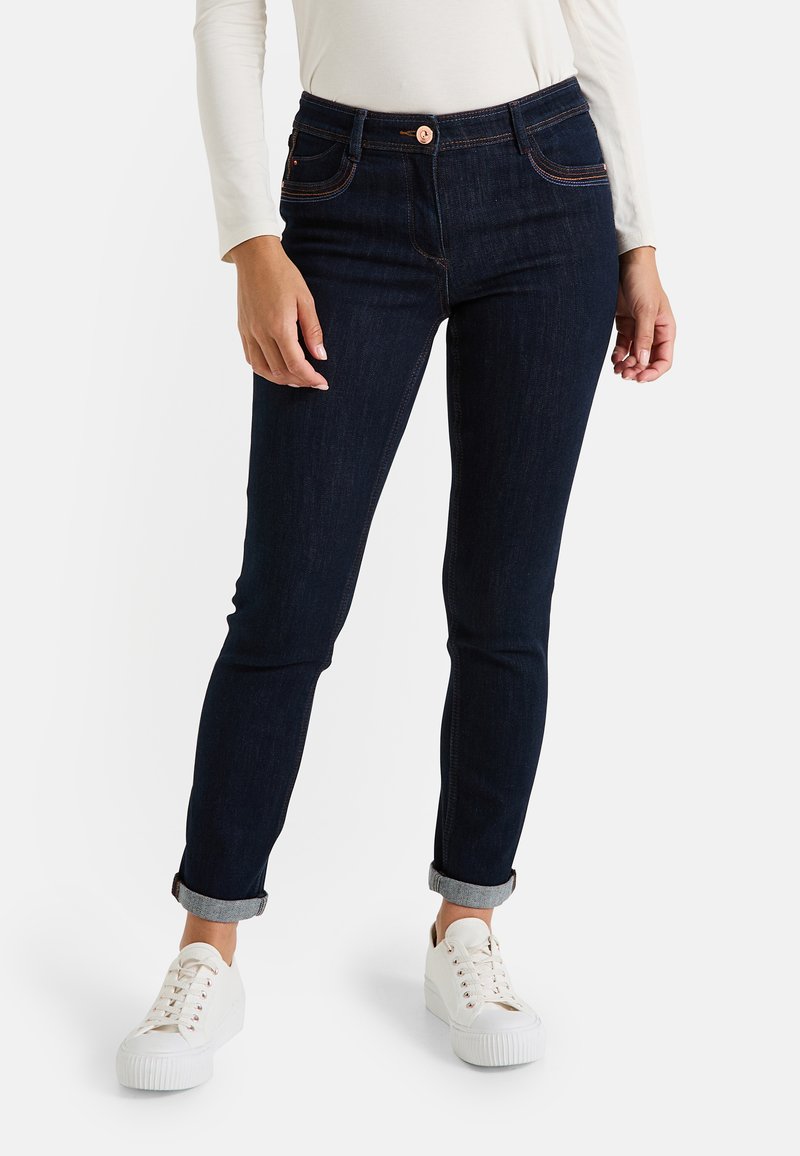 Jean en denim bleu foncé avec une coupe slim, des poignets roulés, un bouton en cuivre et des coutures orange contrastantes. Associé à des baskets blanches.