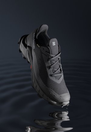 Chaussure de trail Salomon noire avec des gouttes d'eau, flottant au-dessus d'une eau sombre ondulée reflétant l'avant de la chaussure.