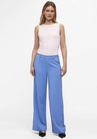 Helles rosa ärmelloses Top, kombiniert mit blauen weiten Hosen mit zwei Fronttaschen, glatter Textur und einer taillierten Passform. Schwarze spitze Schuhe.