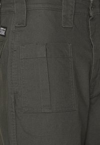 Dunkelgrüne Hose mit seitlicher Cargo-Tasche, aus robustem Material. Die Nähte sind entlang der Taschenränder sichtbar. Minimale Rückenmarkierung vorhanden.