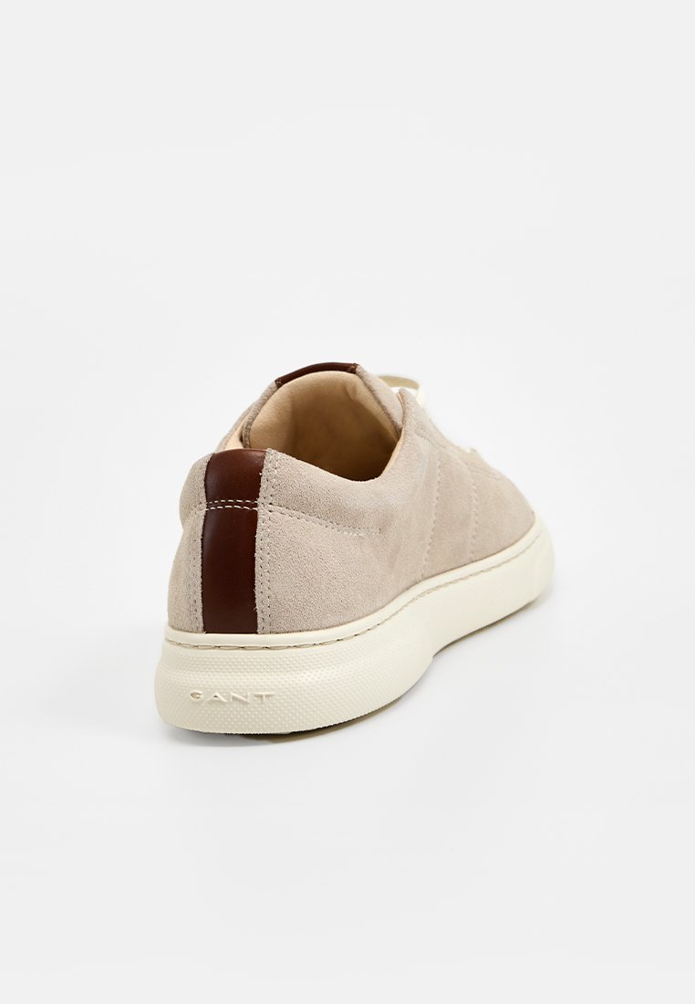 Beige mockasin sneakers med en brun läderdetalj vid hälen och en texturerad vit gummisula. Enkelt designad med rundad form.