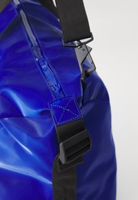 Blue waterproof bag featuring a glossy finish, contrasting black strap, and metallic hardware. Notable detail: blue stitched accents.