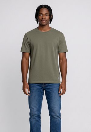 Młody mężczyzna o średnio-ciemnej karnacji i krótkich dredach, ubrany w oliwkową koszulkę i niebieskie jeansy, stojący na tle jasnoszarego tła.