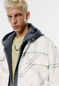 Biely károvaný kabát s šedými akcentmi, vrstvený cez pruhovanú žltú košeľu. Strieborný náhrdelník s detailom v zelenej farbe. Hladká textúra.