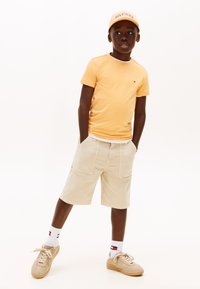 Αγόρι που φοράει ροδακινί καπέλο και μπλουζάκι, μπεζ σορτς, λευκές κάλτσες και μπεζ αθλητικά παπούτσια, στέκεται με τα χέρια στις τσέπες μπροστά σε λευκό φόντο.