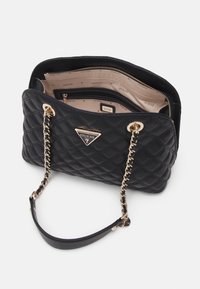 Musta tikattu käsilaukku kultaisen sävyisillä metalliosilla, jossa on ketjuhihna ja tilava sisäpuoli vaaleanbeigellä vuorauksella sekä brändäystiedoilla.