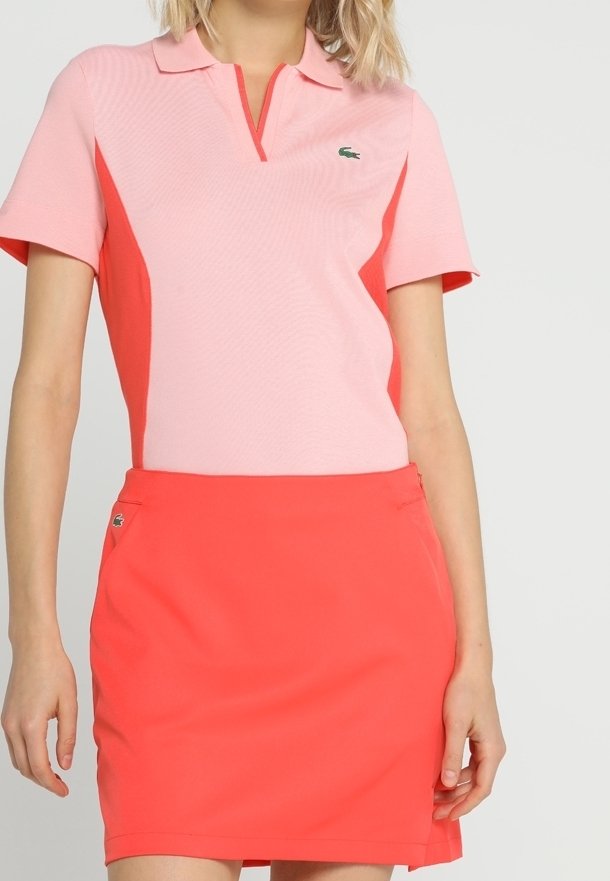 Krage polo i ljusrosa och korall, med korta ärmar och en strukturerad yta, tillsammans med en matchande kjol i korall. Lacoste-logotyp synlig.