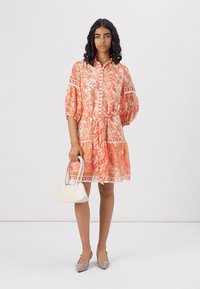 Kjole med korte ærmer i paisley-mønster i orange og pink nuancer, indsnævret i taljen, parret med en hvid håndtaske og beige spidse flade sko.