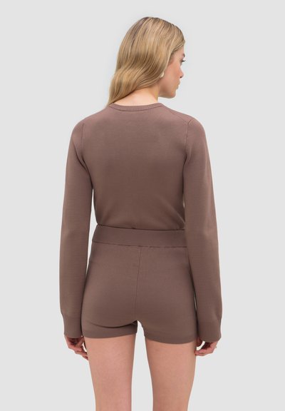 Femme aux cheveux blonds portant un haut marron à manches longues assorti et un short taille haute, de dos sur un fond uni.