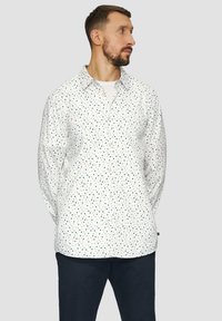 Witte blouse met een stippenpatroon in groen en marineblauw. Voorzien van een klassieke kraag en lange mouwen. Draagt met donkere broek.