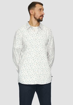 Witte blouse met een stippenpatroon in groen en marineblauw. Voorzien van een klassieke kraag en lange mouwen. Draagt met donkere broek.