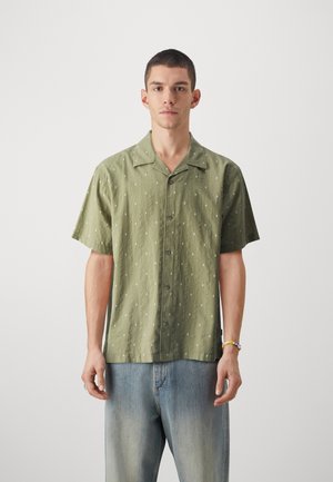 Camisa de manga corta color verde oliva con un patrón de manchas blancas, cierre frontal de botones y un corte holgado, combinada con jeans de color azul claro.