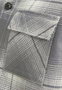 Grau-weißer Karostoff mit einer Klapptasche, detaillierten Nähten und einem schwarzen Vierlochknopf oben links.