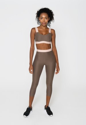 Woman standing wearing brown sports bra and high-waist cropped leggings with beige trim, paired with black and white sneakers, against white background.