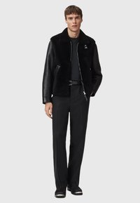 Black shearling and leather jacket with a zip closure, featuring a collar and side pockets, worn over a black top and trousers.