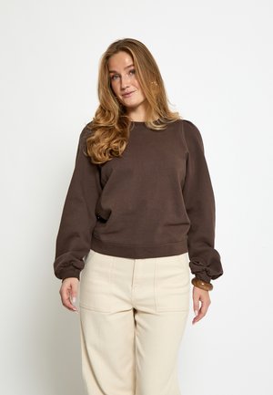 Ung kvinde med langt bølget blondt hår iført en brun langærmet sweatshirt og lysebeige bukser mod en ensfarvet hvid baggrund.