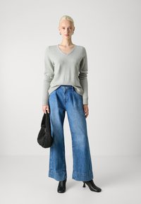 Maglione grigio chiaro a collo a V, jeans larghi in denim blu, stivali neri con tacco. Borsa nera a maglia in mano, tessuto morbido e vestibilità casual.
