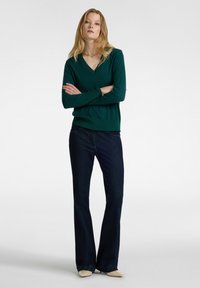 Groene v-hals trui met lange mouwen, verfraaide manchetten, gecombineerd met donkerblauwe flared jeans en lichtgekleurde puntige schoenen.