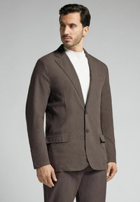 Brauner, einreihiger Blazer aus strukturiertem Stoff, mit zwei vorderen Taschen und Reverskragen. Kombiniert mit einem hellen Oberteil.
