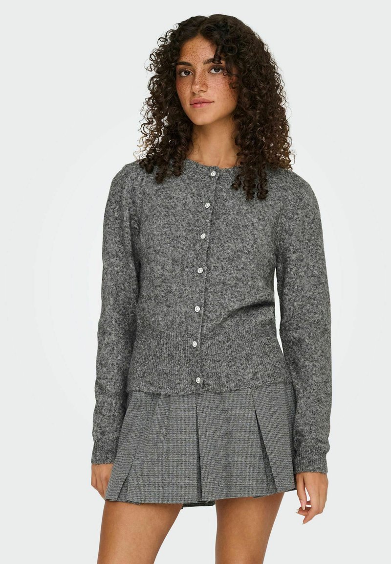 Cardigan tricoté gris avec encolure arrondie et fermeture à boutons, présentant un tissu texturé et des poignets côtelés, associé à une jupe plissée grise.
