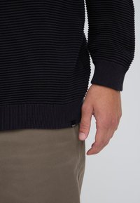 Main d'homme reposant à côté du torse portant un pull noir texturé et un pantalon kaki sur un fond uni.