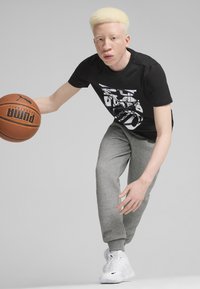 Sort kortærmet T-shirt med hvid grafik, grå sweatpants og hvide sneakers. Basketball i højre hånd. Lys baggrund.