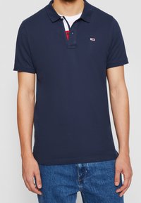 Marinblå polo skjorta i bomull, med krage, två knappslå och en liten logotyp-patch på bröstet. Bärs med denimjeans.