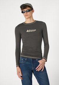 Camiseta gris ajustada de manga larga con un acabado texturizado y costuras de color crema. Presenta un logo estilizado "HYPER" en la parte delantera.