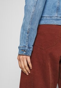 Džínová bunda světle modré barvy s knoflíkovým manžetovým zakončením, v kombinaci s tmavě červeno-hnědými manšestrovými kalhotami s prohnutým švem zadní kapsy.