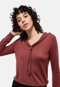 Haut long en maho sur manches avec un col en V et un d�étail noué au niveau du col. Fabriqué en tissu côtelé, il présente une silhouette ajustée et une texture douce.