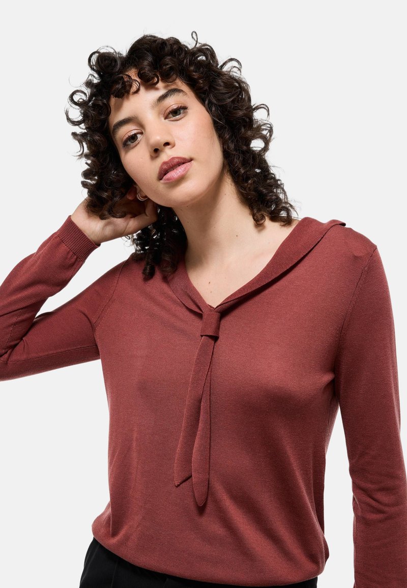 Haut long en maho sur manches avec un col en V et un détail noué au niveau du col. Fabriqué en tissu côtelé, il présente une silhouette ajustée et une texture douce.