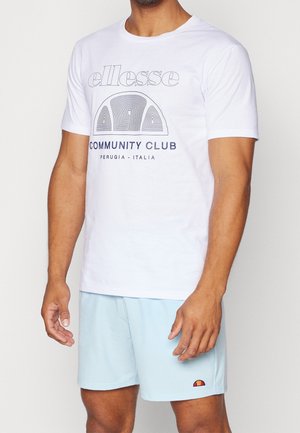 Mand iført hvid t-shirt med teksten "ellesse Community Club Perugia - Italia" og lyseblå shorts med lille logo på venstre kant.
