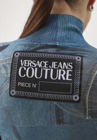 Mėlynas džinsinis drabužis su raukšlėtu apykakle, turintis didelį juodą etiketę su užrašu "VERSACE JEANS COUTURE", taip pat su tuščiu lauku "PIECE N°."