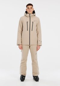 Beige skijack en -broek van glad, waterdicht materiaal. De jack heeft een capuchon, zakken met ritsen en zwarte accenten.