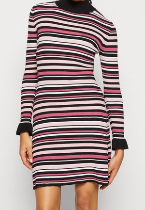Frau trägt ein eng anliegendes, geripptes Strickkleid mit horizontalen Streifen in Schwarz, Weiß, Pink und Hellpink, mit einem schwarzen Rollkragen und Rüschen an den Ärmelenden.