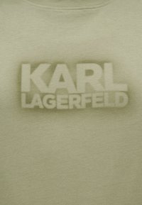 Tissu beige avec le texte « KARL LAGERFELD » en grandes lettres grasses légèrement floues centrées sur la surface.