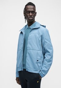 Casaco corta-vento azul-claro com fecho de correr frontal, capuz e dois bolsos laterais. Usado sobre uma camisa pólo verde-petróleo e combinado com calças escuras.