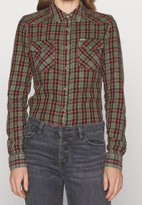 Camicia a quadri con bottoni in verde, rosso e nero. Presenta due tasche sul petto, maniche lunghe e un paio di jeans grigi scuri.
