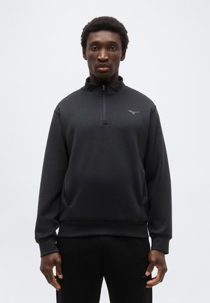 Άνδρας που φοράει μαύρο πουλόβερ με μακριά μανίκια, με φερμουάρ στο ένα τέταρτο και μικρό λογότυπο στο αριστερό στήθος, στέκεται μπροστά σε απλό φόντο.