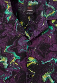 Chemise à manches courtes boutonnée en noir, présentant un motif marbré de violet, vert et jaune. Comprend un col plié et des boutons noirs.