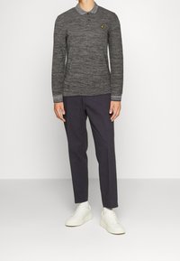 Homme portant un polo à manches longues gris foncé, un pantalon noir slim, des chaussettes blanches et des baskets blanches se tenant devant un fond uni.