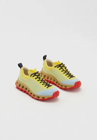 Yellow and blue mesh sneakers with a brown midsole and red sole. Features black laces and a rear pull tab. Textured surface detail.