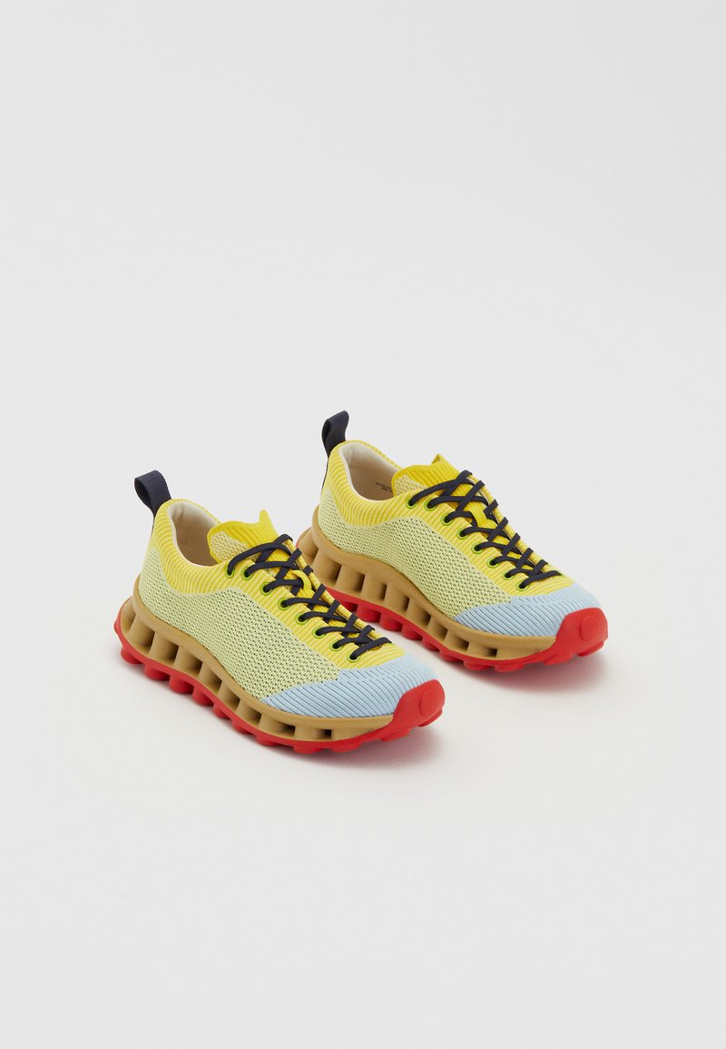 Yellow and blue mesh sneakers with a brown midsole and red sole. Features black laces and a rear pull tab. Textured surface detail.
